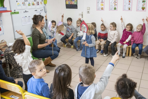 Classe de maternelle de Notre-Dame-de-Lourdes, les enfants sont assisen cercles autour de la maîtresse et une petite a un masque dessiné
